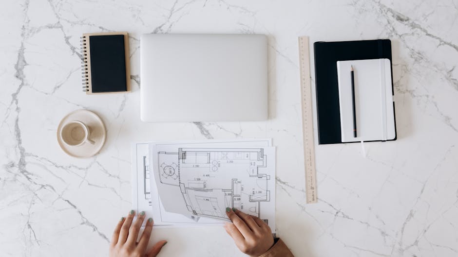 Top view of an architect's desk featuring blueprints, laptop, notebook, and coffee cup on a marble surface.